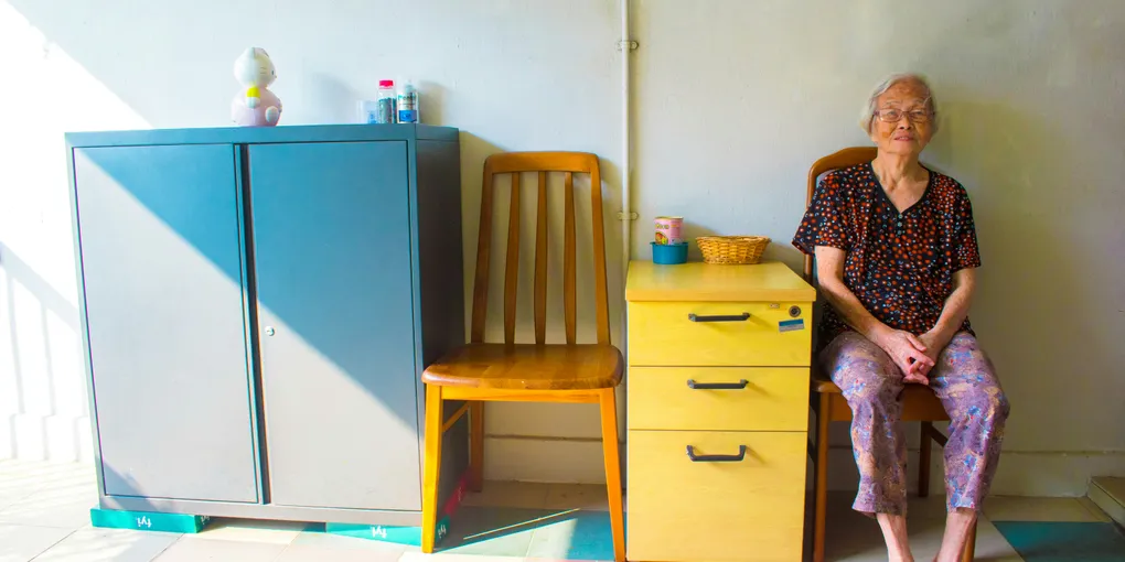 Femme âgée assise dans une chambre claire, à côté d'une chaise vide et d'une petite commode